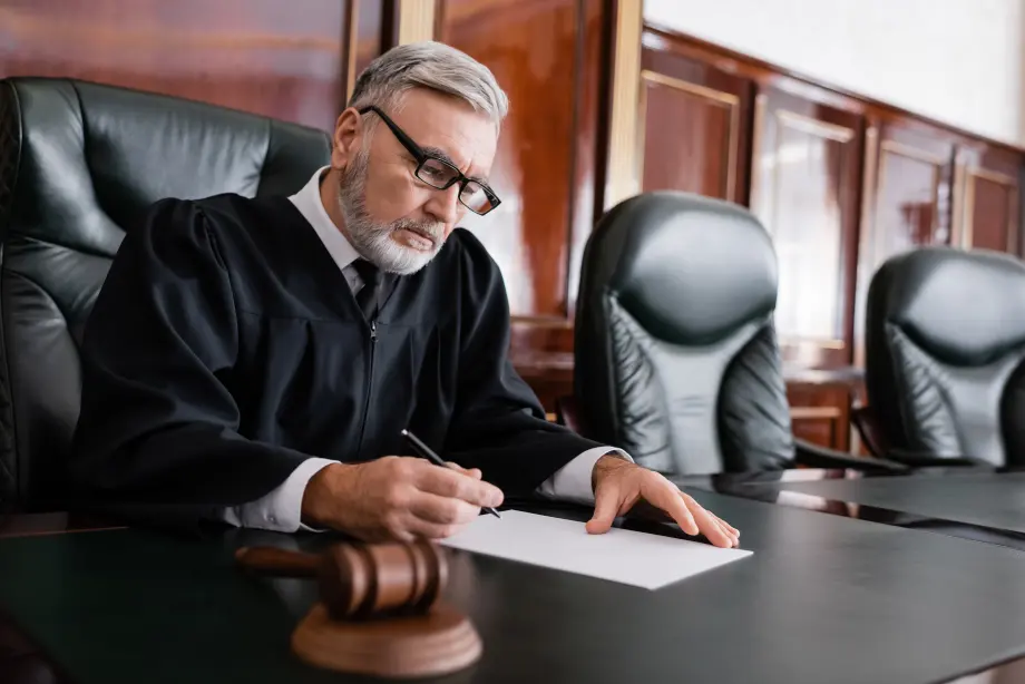 A judge with gray hair and glasses writes on a document at a desk in a courtroom, with a gavel nearby.