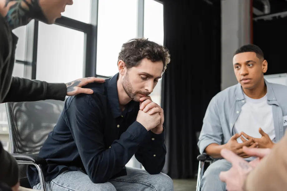 A man sits with his head down, appearing thoughtful or distressed, while others around him offer support in a group setting.