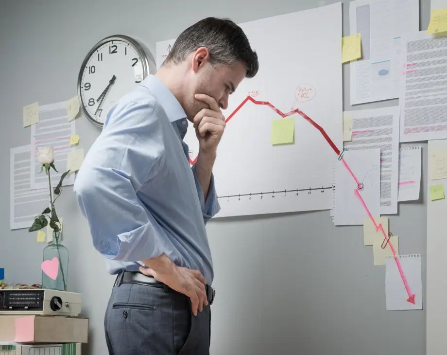 A man stands pensively in front of a wall chart displaying a steep downward trend, surrounded by notes and documents, with a clock and a rose in the background.