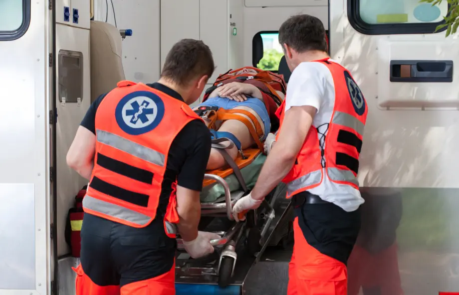 Two paramedics in orange vests load a patient on a stretcher into the back of an ambulance.