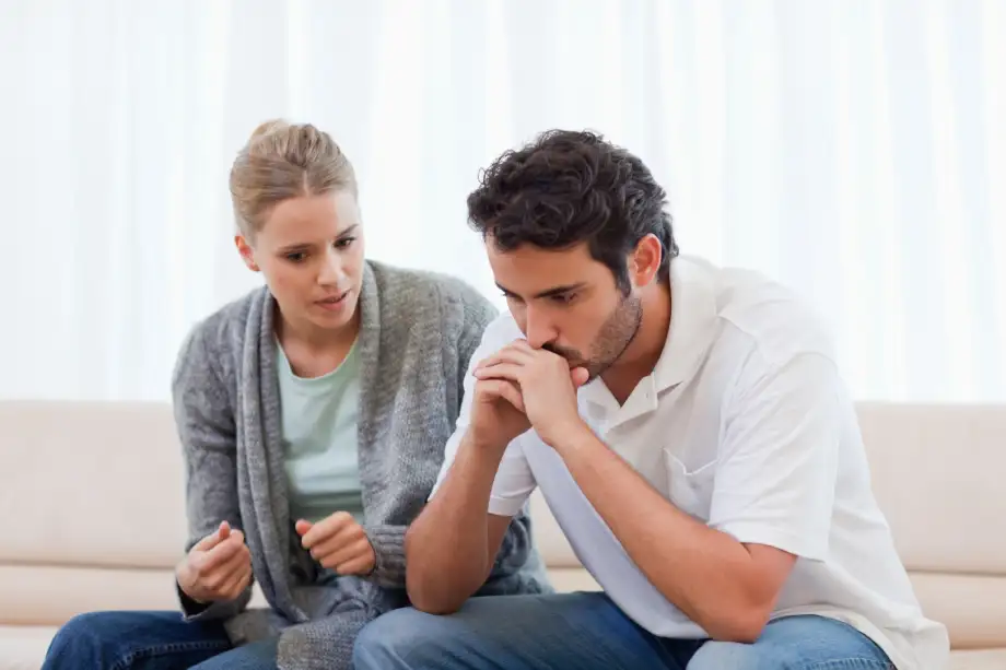 A woman sitting on a couch appears to be talking to a man beside her, who is leaning forward with his hands near his mouth, looking thoughtful or concerned during an estate plan goals discussion.