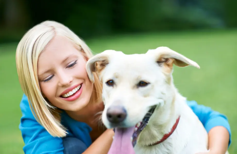 A woman with blonde hair smiles while hugging a large, light-colored dog outdoors on a grassy area.