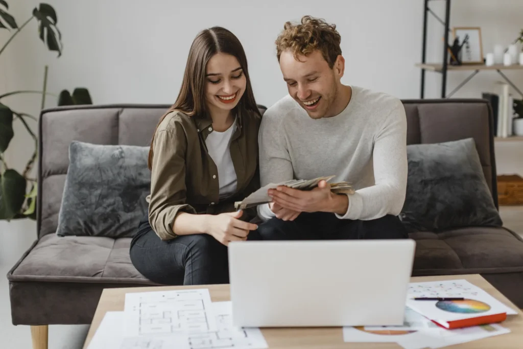 A man and woman sit on a sofa, smiling while looking at papers in front of an open laptop on a coffee table with documents and a color wheel, discussing ways to avoid wire transfer fraud in real estate.