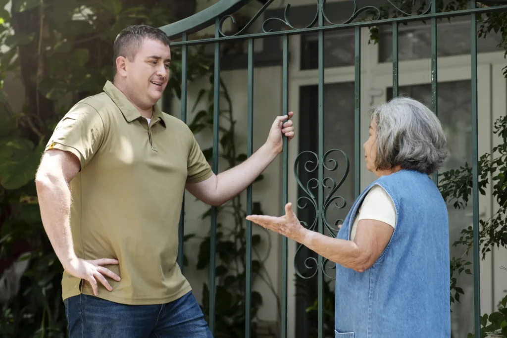 A man in a tan polo shirt discusses boundary disputes between neighbors with an older woman in a blue vest by a metal gate outside a house.