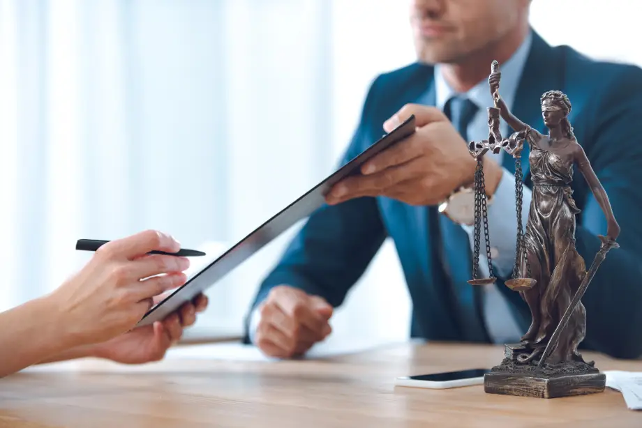 Two people sit at a desk, one handing a clipboard to the other. A bronze Lady Justice statue is on the table in the foreground.