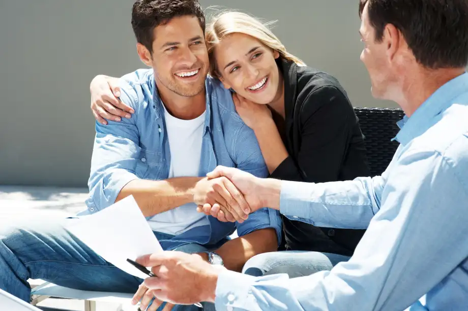 A couple sits together, smiling, as the man shakes hands with another man holding paperwork during an estate planning for business owners consultation.