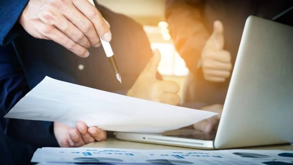 Two people review essential estate planning documents and charts near a laptop; one holds a pen and paper while another gives a thumbs up, suggesting agreement or approval.