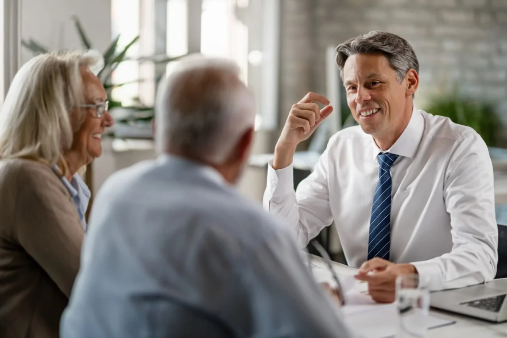 A man in a suit is smiling and talking with two older adults across a table in a bright office setting, discussing ways to protect your assets.