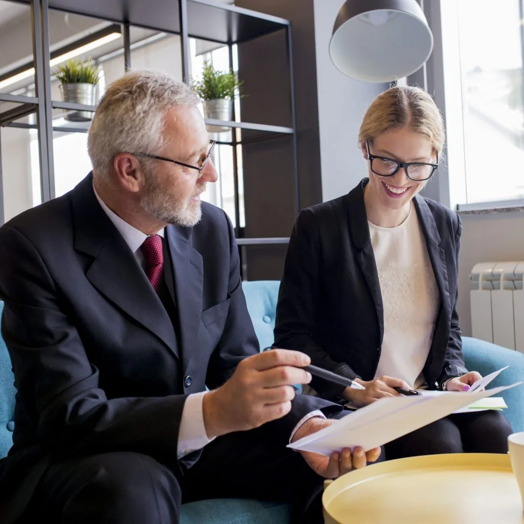 Two people in business attire sit on a couch, reviewing documents about Trusts for Protection and Distribution together in a modern office setting.