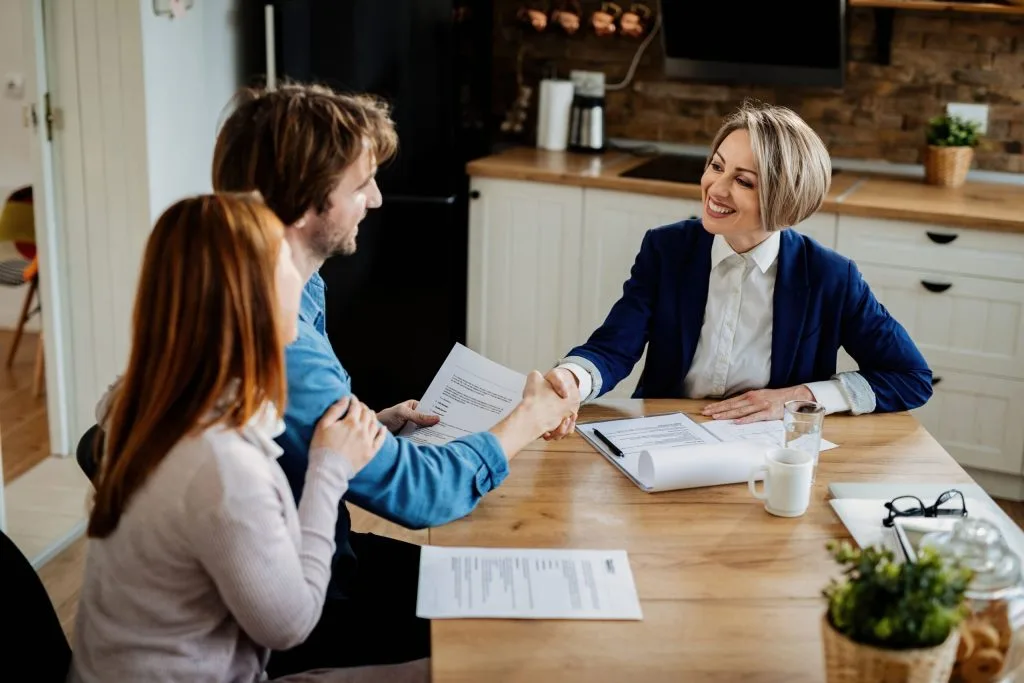 A couple sits at a table with a woman in business attire, shaking hands and holding documents, suggesting a meeting to discuss how to avoid probate in South Carolina in a modern kitchen setting.