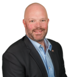 A bald man with a beard wearing a dark suit jacket and light blue shirt smiles at the camera against a white background.