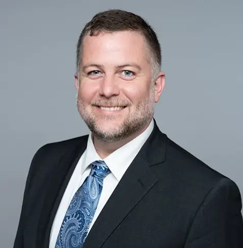 Harrison Long of Sawyer & Associates, dressed in a dark suit, white shirt, and blue patterned tie, smiles at the camera against a plain grey background.