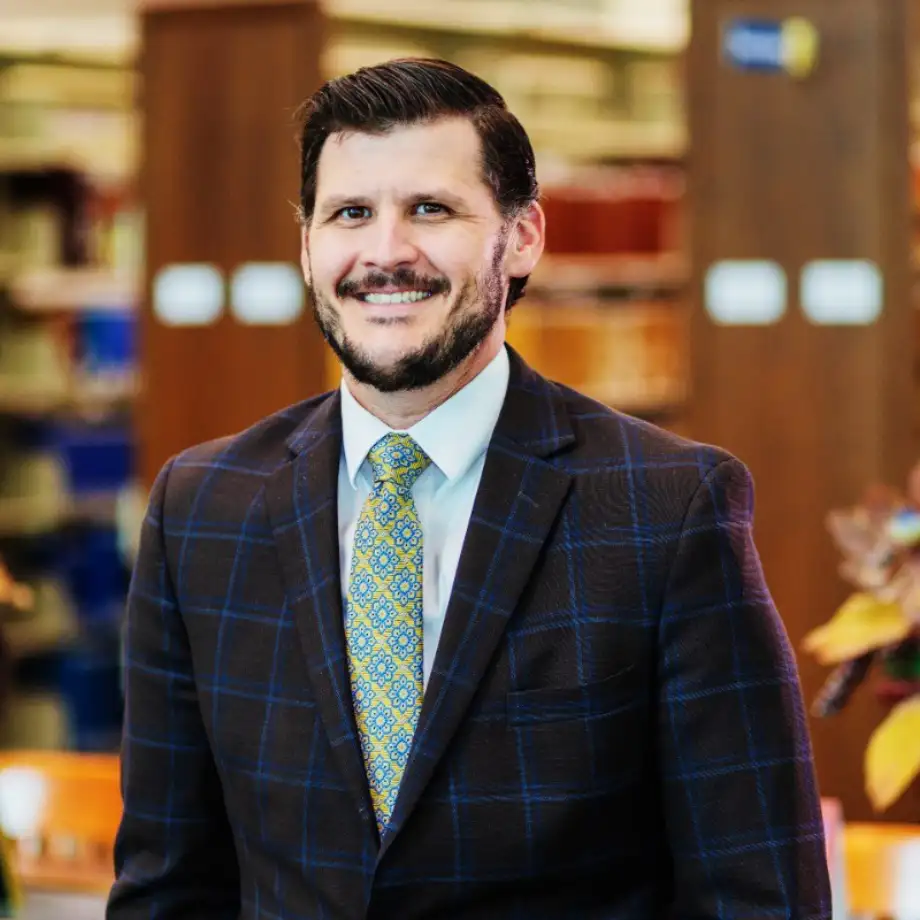 Sawyer & Associates stands indoors in a dark checked suit, light shirt, and patterned tie, smiling with bookshelves and plants in the background.