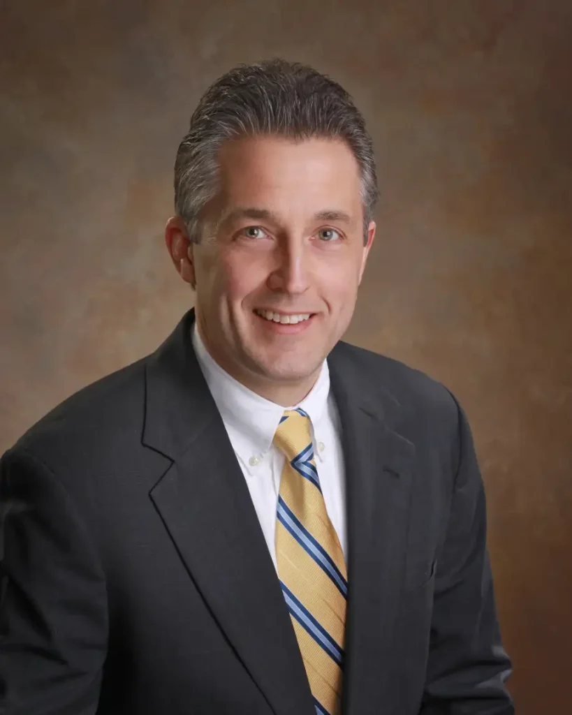 Matt Lindauer of Sawyer & Associates sits in front of a brown studio backdrop, smiling at the camera in a dark suit, white shirt, and striped yellow tie.