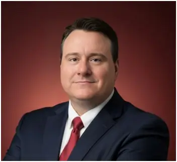 A man from Nick Wilson Sawyer & Associates, dressed in a dark pinstripe suit, white shirt, and red patterned tie, stands confidently before a plain gray background, looking directly at the camera.