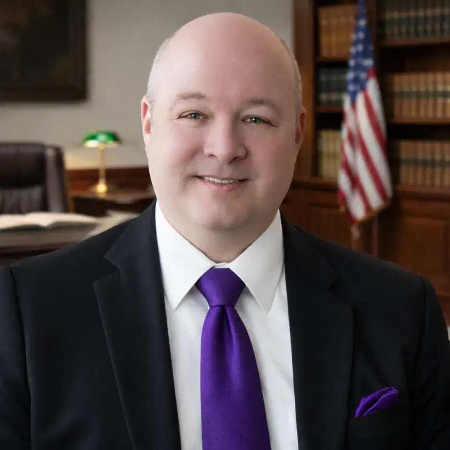 Rob Schaefer of Sawyer & Associates, dressed in a suit and purple tie, sits in an office with bookshelves, a desk, and an American flag in the background.