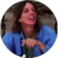 A person with long hair, wearing a blue shirt, sits outdoors and appears to be laughing or speaking. The image is slightly blurred.