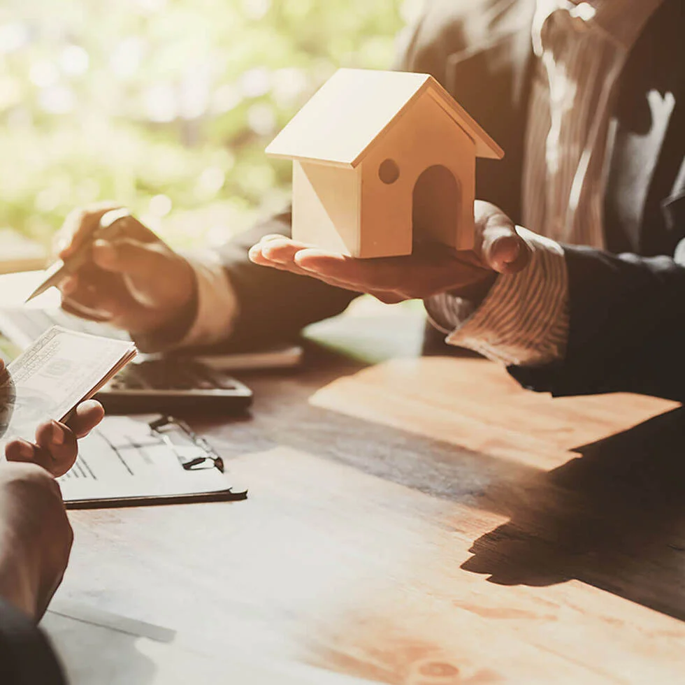 Two people in business attire sit at a table with documents, one holding a model house and the other holding a pen and a checkbook, suggesting a real estate transaction.